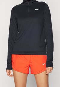 Černé sportovní tričko s dlouhým rukávem a čtvrtinovým zipem, na kterém je logo Nike, spárované s oranžovými sportovními šortkami s zakřiveným lemem a logem Nike.