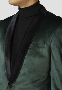 Grüner Samtblazer mit schwarzem Satin-Schalkragen, Ein-Knopf-Verschluss und dezentem Glanz, kombiniert mit einem enganliegenden schwarzen Rollkragenpullover.