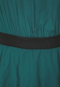 Türkisfarbener Stoff mit glatter Textur, der sich an der Taille sammelt und ein breites schwarzes Elastikband aufweist. Einfaches Design, keine Muster oder zusätzlichen Details.