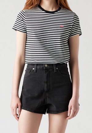 Osoba nosząca czarno-biały pasiasty t-shirt Levi's włożony do czarnych, wysokich szortów jeansowych na jednolitym tle.