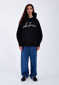 Sort hættetrøje med snore og stor hvid tekst på fronten. Kombineret med løstsiddende blå denimjeans og sorte sko, stående på en hvid baggrund.