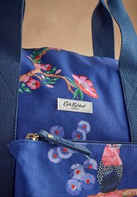 Mėlyna drobės pirkinių maišelis su gėlių raštais, užtrauktuku užeiga ir etikete, kurioje parašyta "Cath Kidston". Rankenos tamsiai mėlynos ir plokščios.