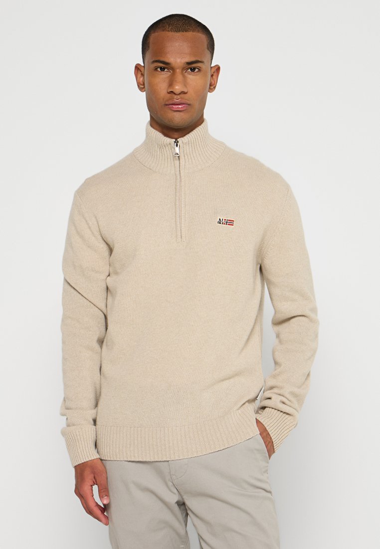 Un homme portant un pull à demi-fermeture éclair beige avec un petit logo drapeau sur la poitrine et un pantalon beige clair, se tenant devant un fond blanc uni.