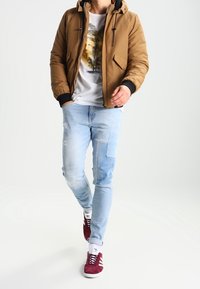 Brown padded jacket with hood, worn over a white t-shirt featuring a palm tree graphic. Light blue distressed jeans and burgundy sneakers.