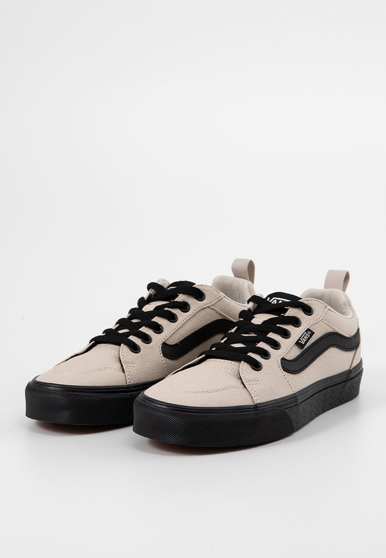 Une paire de baskets en toile beige avec des lacets noirs, des semelles noires et une bande latérale Vans noire, présentée sur fond blanc.