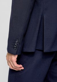 Ärmel einer marineblauen Anzugjacke aus strukturiertem Stoff mit drei schwarzen Knöpfen und einem sichtbaren Schlitz auf der Rückseite, auf dem eine Hand ruht.