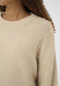 Maglione lavorato a maglia in beige chiaro con un colletto a coste. Perle decorative di colore crema sparse sulle spalle e sulle maniche. Tessuto morbido.