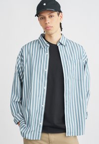 Pruhovaná košeľa v svetlomodie a bielej farbe z bavlny, zapínanie na gombíky s jednou náprsní prackou. Nosená cez čierne tričko a v kombinácii s khaki nohavicami.