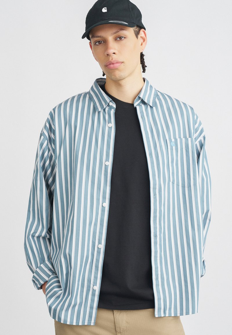 Pasiasta koszula w jasnoniebieskim i białym kolorze z bawełny, zapinana na guziki, z jedną kieszenią na piersi. Nosi się ją na czarną koszulkę i w połączeniu z beżowymi spodniami.