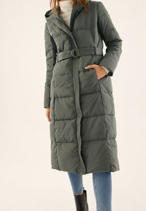 Femme portant un long manteau matelassé vert ceinturé avec une capuche, un col roulé blanc, un jean bleu et des bottines noires, debout devant un fond uni.