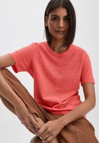 Korallinvärinen lyhythihainen t-paita pehmeästä, kevyestä kankaasta, pyöreällä kaula-aukolla, yhdistettynä ruskeisiin löysiin housuihin. Huomaamaton tekstuuri näkyvissä.