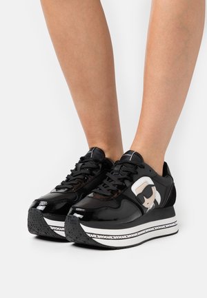 Sneaker in pelle verniciata nera con un design di cartone bianco e beige sul lato, suola intermedia spessa bianca e suola esterna nera testurizzata.