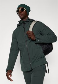 Donkergroene waterdichte jas met ritssluiting en zakken, gecombineerd met bijpassende broek, een groene beanie en oversized lichtgekleurde zonnebril.