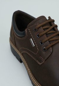 Zapato de piel marrón con cordones, costuras de contraste, ojales de metal, un cuello acolchado negro y una etiqueta metálica de la marca en el costado.