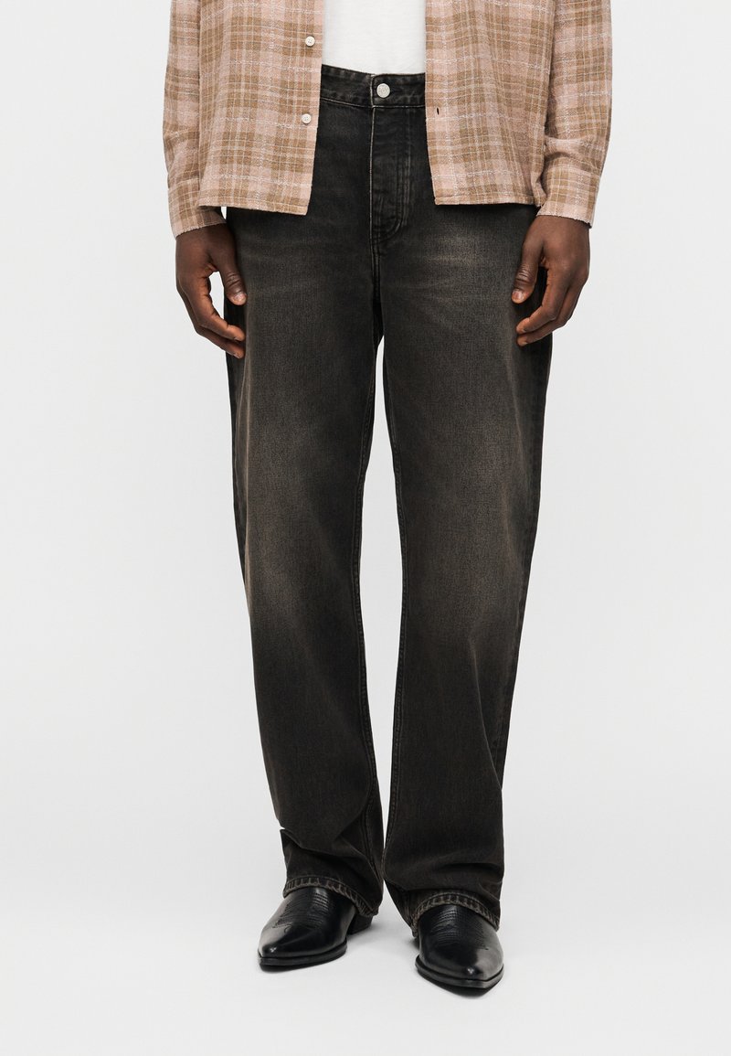 Κάτω μέρος σώματος ενός ατόμου που φοράει ξεθωριασμένα μαύρα τζιν φαρδιάς γραμμής, μαύρα μυτερά παπούτσια και μια μπεζ πουκάμισο καρό ορατό στη μέση.