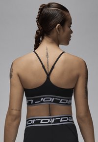 Sujetador deportivo negro con tirantes ajustables y una banda elástica ancha que presenta el logo "JORDAN" en gris. Textura suave y diseño ajustado.