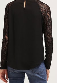 Blusa nera con maniche lunghe in pizzo, dettaglio a occhio di keyhole sul retro e orlo curvo. Tessuto morbido per il corpo, dettagli in pizzo testurizzato sulle maniche.