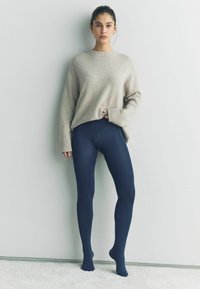 Světle béžový oversized svetr s žebrovan�ými manžetami v kombinaci s tmavě modrými legínami. Legíny mají bezešvý design a průsvitnou texturu.