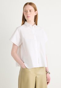 Hvit kortermet skjorte med knapper og krage, avslappet passform og glatt bomullsstoff. Kombinert med lyse beige bukser.