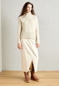 Krem strikkegenser med ribbestrikket kant, parret med en beige midikjole med frontsliss; brune snørebots med pelsdetaljer.