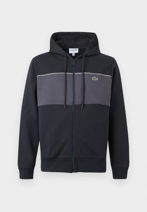 Μαύρο και σκούρο γκρι φούτερ με φερμουάρ, κουκούλα με κορδόνια και λογότυπο πράσινου κροκόδειλου Lacoste στο στήθος.