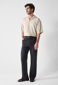 Kurzärmeliges cremefarbenes Hemd mit Knöpfen und gerippter Textur, kombiniert mit schwarzen geraden Hosen und schwarzen Riemchensandalen.