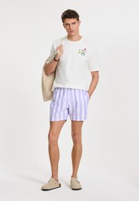 Fehér pamut póló grafikai nyomattal, világos lila és fehér csíkos fürdőnadrággal párosítva. Woven táskát tart, bézs slip-on cipő van rajta.