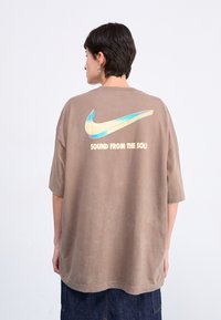 Brun oversized t-shirt lavet af blødt stof, med et stort Nike-logo i blå og guld på ryggen, samt teksten "SOUND FROM THE SOLE."