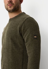 Mørkegrøn strikket sweater med ribbet tekstur på siderne og rund halsudskæring. Har et lille sort logo-patch på venstre ærme.