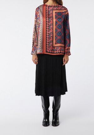 Nainen, jolla on monivärinen kuvioitu pitkähihainen pusero, musta laskostettu midi-hame ja mustat polven yli ulottuvat nahkasaappaat, seisomassa valkoista taustaa vasten.