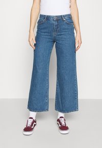 Blå vida jeans med hög midja, med framfickor och knappstängning. Bärs med bordeauxfärgade sneakers och vita strumpor.