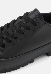 Baskets en cuir noir avec semelle en caoutchouc épaisse, lacets noirs passés à travers des œillets métalliques, et nom de la marque discrètement embossé sur la languette.