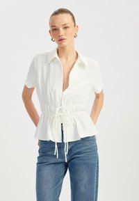 Hvid skjorte med knapper, der har et struktureret mønster, korte ærmer og en indsnævret talje, parret med lyseblå denim jeans.