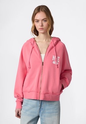 Giovane donna che indossa una felpa rosa con zip e scritte bianche sul petto, jeans denim azzurri chiari, in piedi davanti a uno sfondo bianco.