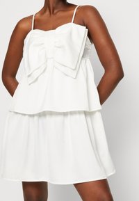 Robe blanche sans manches avec un grand détail de nœud central. Design en couches avec une jupe fluide, fabriquée à partir d'un tissu doux et léger.