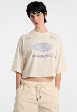 Beige cotton crop t-shirt with relaxed fit, featuring blue and red printed logos. Short sleeves, round neckline, and minimal stitching details.