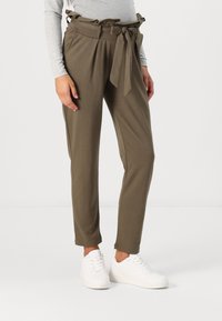 Pantalons vert olive avec une taille à volants et un cordon de serrage, des jambes fuselées et des poches latérales, associés à des baskets blanches sur un fond neutre.
