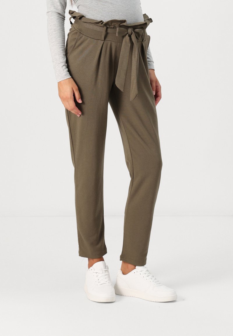 Pantalons vert olive avec une taille à volants et un cordon de serrage, des jambes fuselées et des poches latérales, associés à des baskets blanches sur un fond neutre.