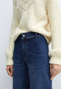 Roomkleurige gebreide trui met een gestructureerd, geribbeld patroon en opgerolde mouwen, gecombineerd met donkerblauwe spijkerbroek met knoopsluiting.