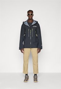 Zwarte waterdichte jas met grijze voering, voorzien van ritszakken en gele accenten, gecombineerd met beige broeken en grijze sneakers.