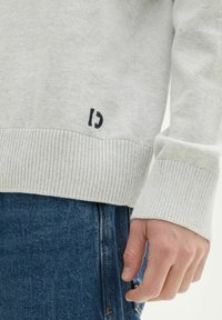Helles graues Sweatshirt mit gerippten Bündchen und Saum, mit einem kleinen dunklen Logodetail. Tragen mit blauen Denim-Jeans, die die Struktur des Stoffs hervorheben.