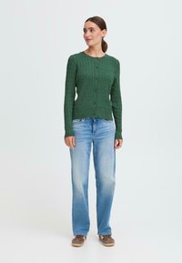 Cardigan en tricot vert avec un motif texturé, col rond et boutons sur le devant, associé à un jean bleu clair à jambes droites et des chaussures marron.