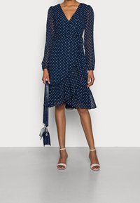 Laivansinis pilkšvas taškelis, su dviem ilgais permatomais rankovėmis, banguotu apačios kraštu ir giliu V formos iškirpimu. Suderinta su maža laivansine tekstūruota rankine ir nuogais aukštakulniais.