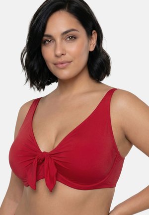 Donna con capelli corti e scuri che indossa un top bikini rosso con un dettaglio nodo frontale, che posa su uno sfondo semplice.