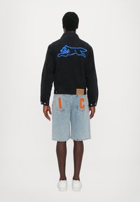 Veste en denim noire avec une broderie de chat bleu en train de sauter au dos, associée à un short en denim bleu clair arborant l'inscription orange "I C." Baskets blanches.