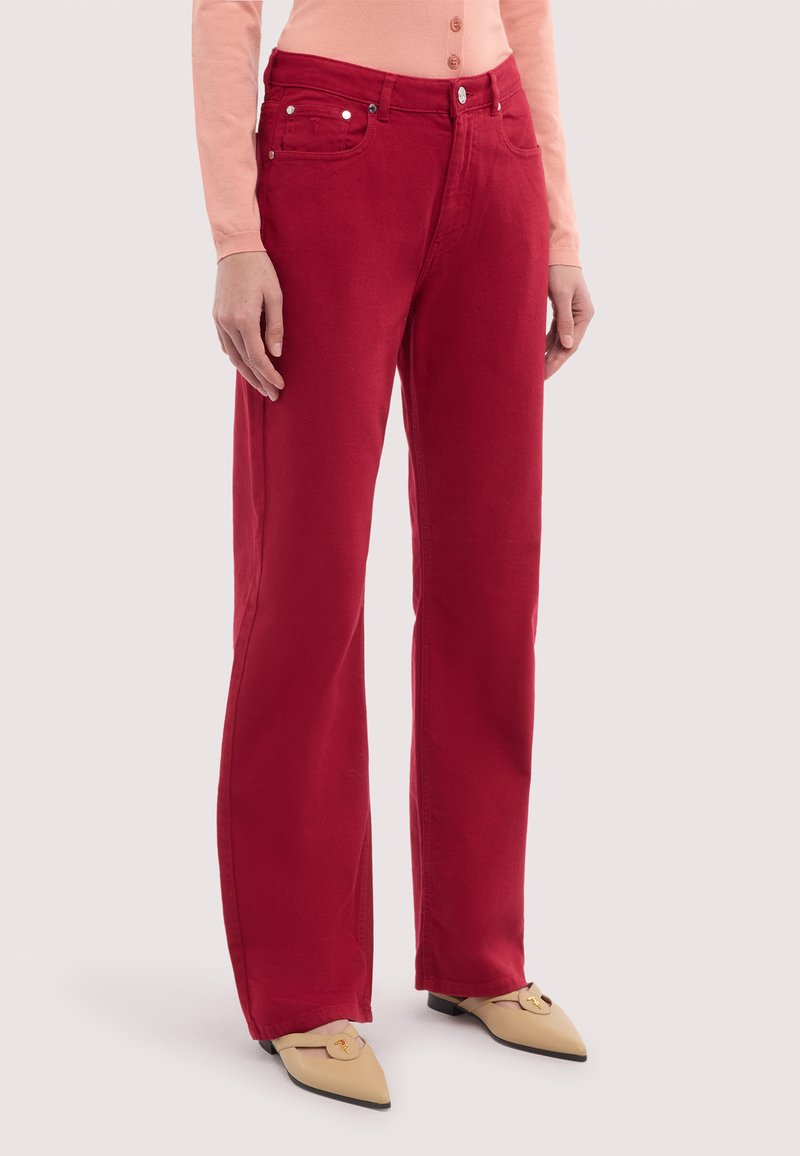 Kobieta ubrana w czerwone, proste spodnie z wysokim stanem, zestawione z beżowymi butami w szpic oraz brzoskwiniową bluzką z długim rękawem.