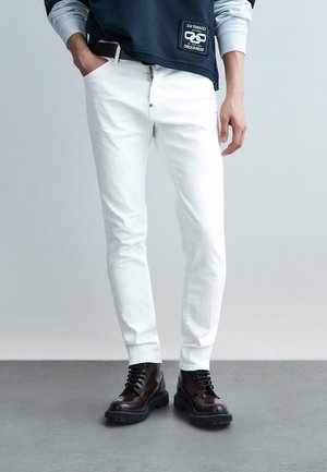 Persoană purtând blugi albi, mulați, cu toc, cizme negre de piele cu șireturi și un hanorac negru cu un patch cu logo, stând pe un podea gri.