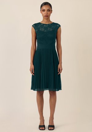 Frau stehend, trägt ein dunkelgrünes ärmelloses Spitzenkleid mit Plisseerock und schwarzen offenen Pumps, neutraler beiger Hintergrund.