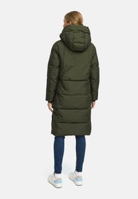 Parka lunga, ampia e di un verde scuro con cappuccio, caratterizzata da un design trapuntato e una silhouette dritta. Indossata sopra jeans blu e sneakers bianche.