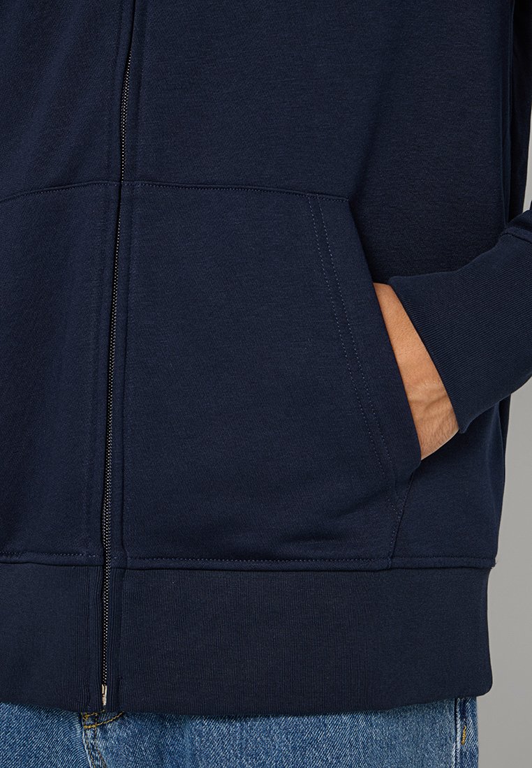 Marinblå zip-up hoodie med en texturerad yta, kängurufickor och ribbade ärmslut. Har en slät dragkedja längs framseam.